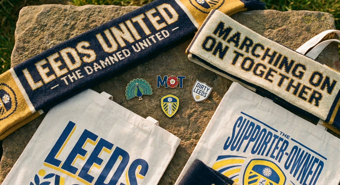 A warm flat lay of independent Leeds United fan merchandise including scarves, enamel pins, and tote bags arranged on a rustic Yorkshire stone surface in golden light.